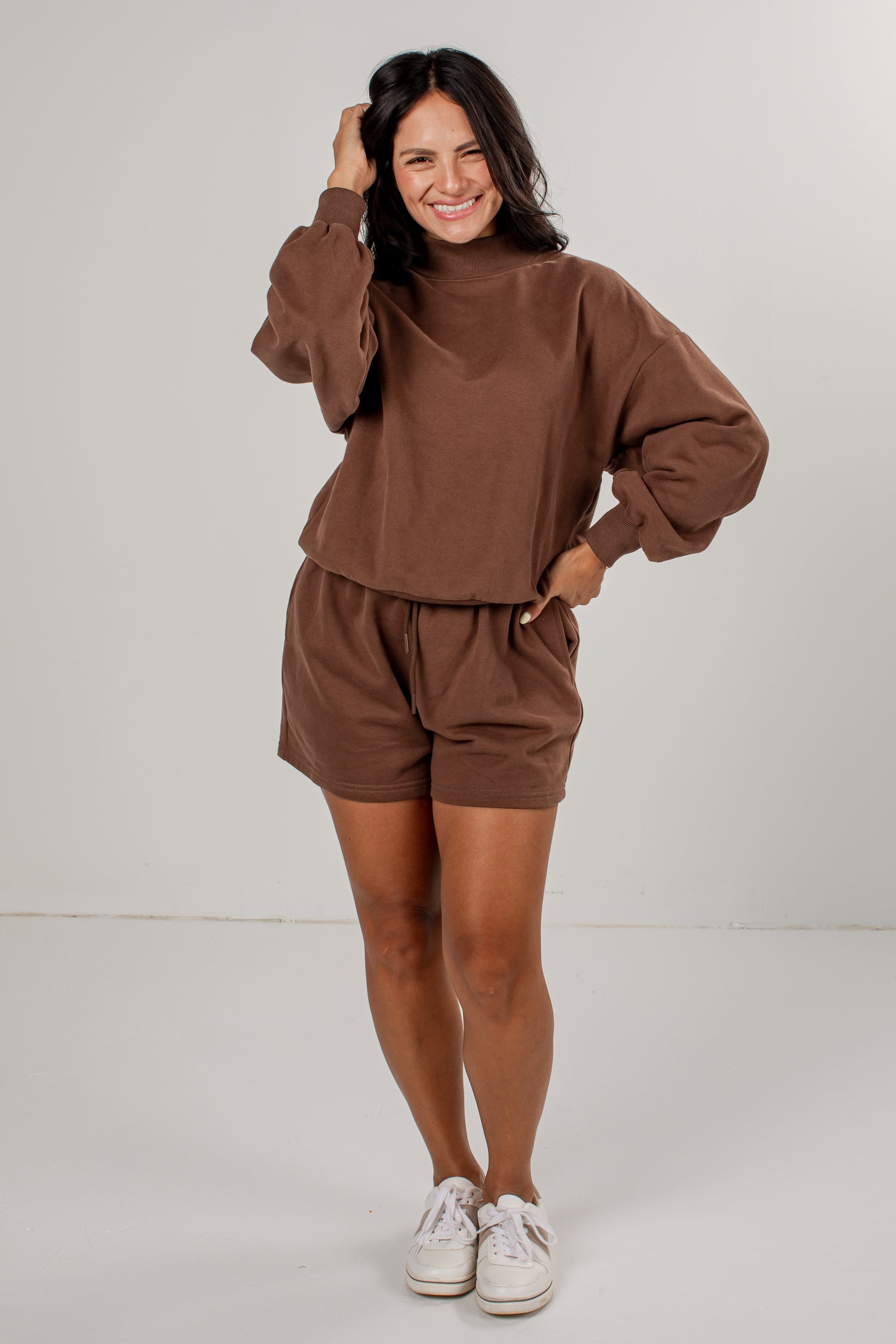 A woman modeling a matching brown lounge set consisting of a long-sleeve mock-neck sweatshirt and drawstring shorts, paired with white sneakers, against a light gray studio background.