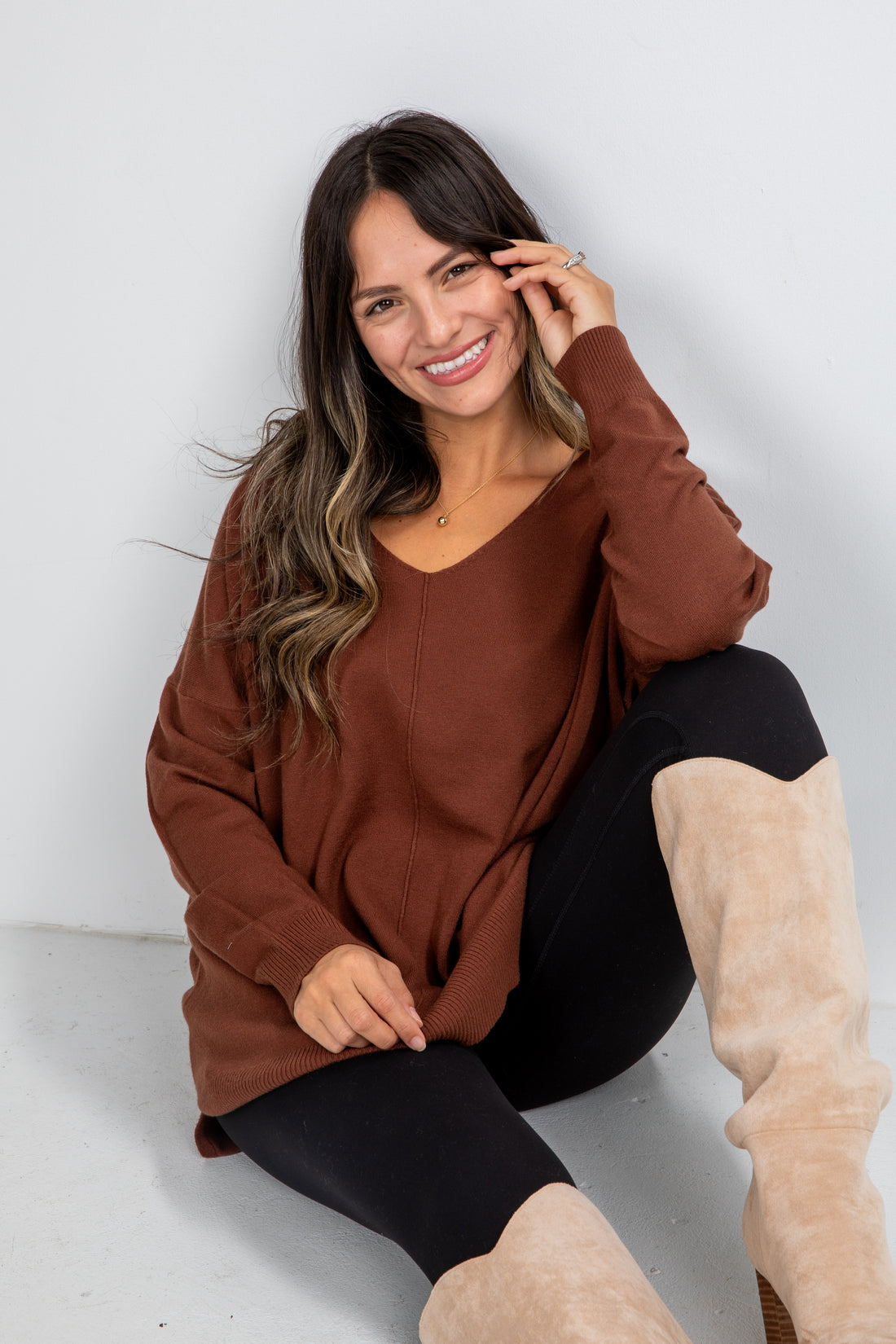 Woman wearing a brown sweater and black leggings with beige boots sitting on a white floor.