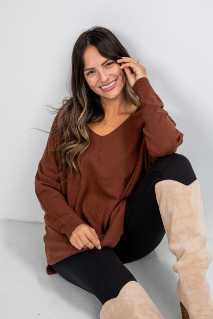 Woman wearing a brown sweater and black leggings with beige boots sitting on a white floor.