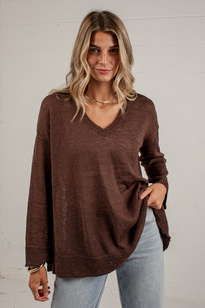 A woman wearing a brown V-neck knit sweater with long sleeves and side pockets, paired with light blue jeans, standing against a white brick wall.