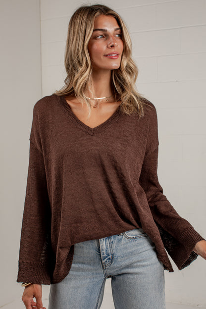 A woman wearing a brown V-neck knit sweater with long bell sleeves and light blue jeans, standing against a white paneled wall.