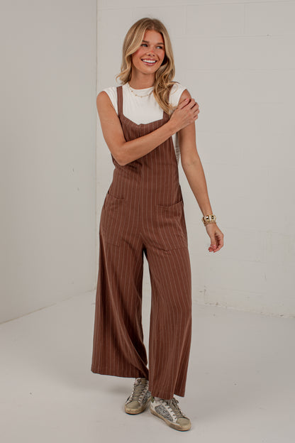 A woman modeling a pair of brown pinstriped wide-leg overalls over a white sleeveless top, standing against a white studio background.