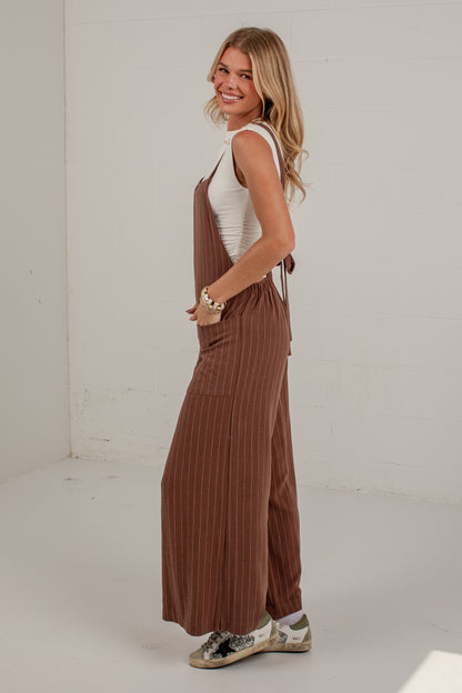 A woman modeling a pair of brown pinstriped wide-leg jumpsuits over a white tank top, standing in a bright white studio.