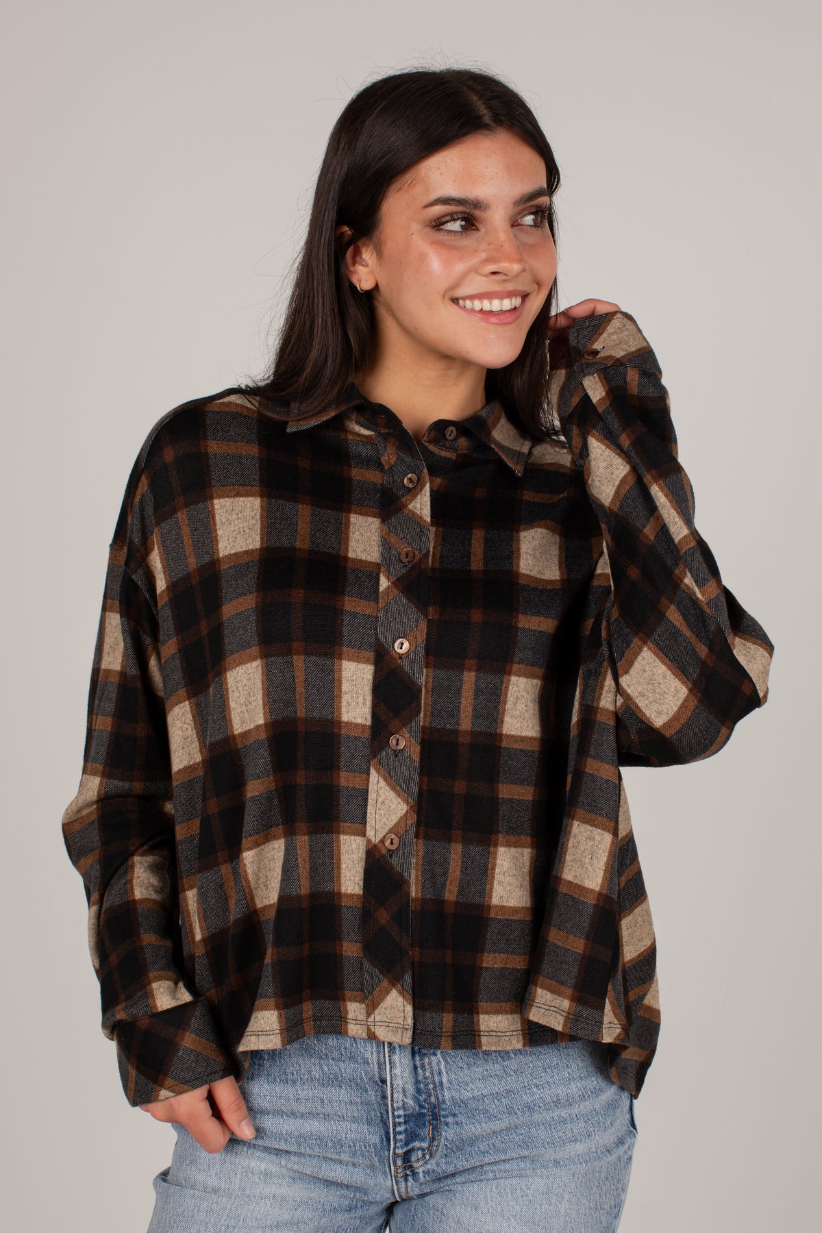 A woman wearing a long-sleeve button-up plaid shirt in brown, black, and beige, paired with light blue jeans, against a solid light gray background.