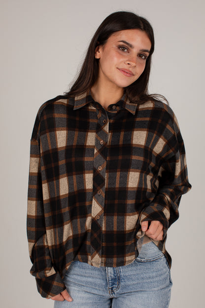 A woman wearing an oversized long-sleeve plaid button-up shirt in brown, black, and beige, paired with light blue jeans, against a plain light gray studio background.