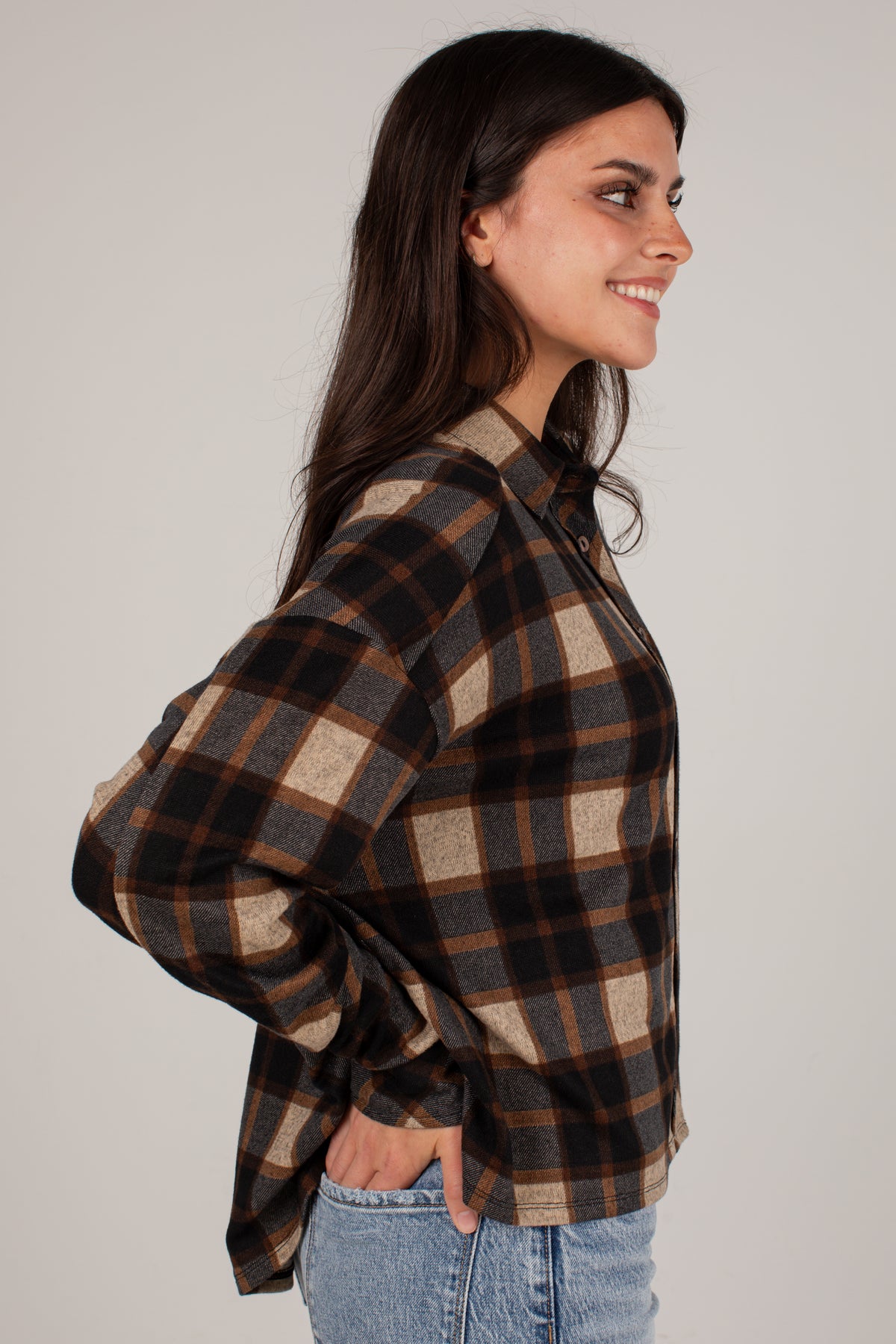 A woman wearing a brown, black, and beige plaid button-up shirt, shown in a side profile view against a plain light gray studio background.