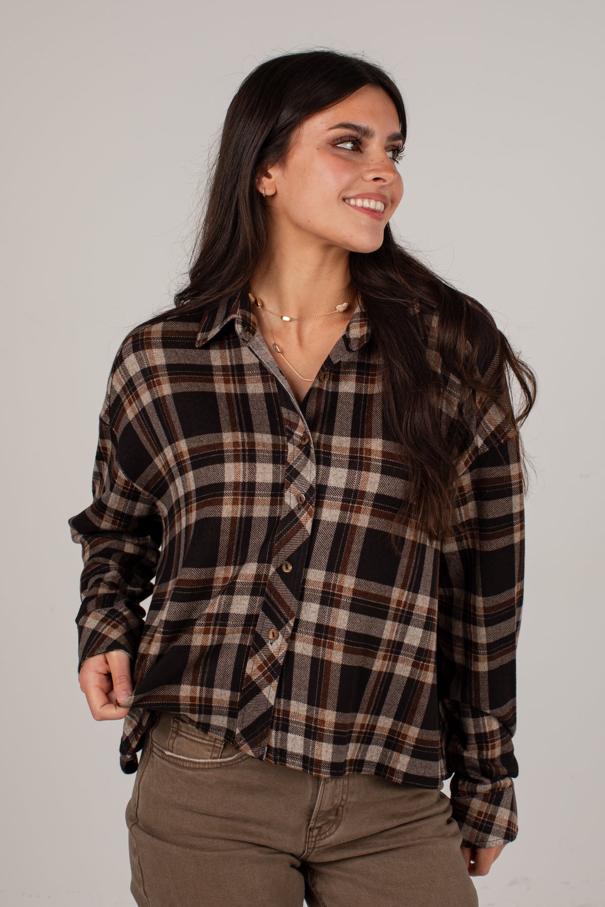 A woman wearing a brown, black, and beige plaid button-up shirt and brown pants, posing against a light gray studio background.