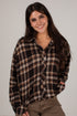 A woman with long dark hair smiling while wearing a brown, black, and beige plaid button-up shirt and brown pants, photographed against a plain light gray background.