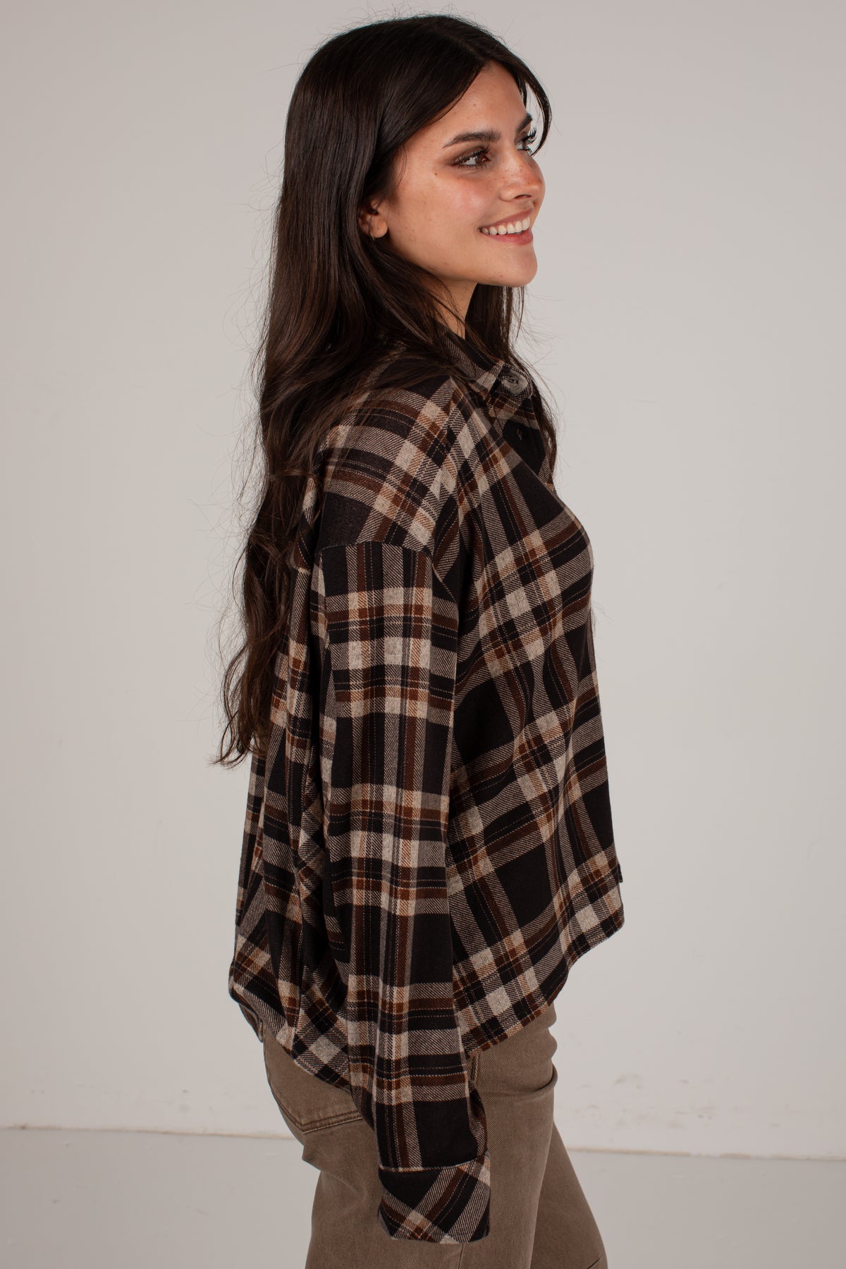 A woman with long dark hair is shown from the side, wearing an oversized brown and black plaid flannel shirt with tan and brown stripes, paired with khaki pants, against a plain off-white background.
