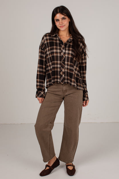 A woman modeling a long-sleeve brown and black plaid button-down shirt paired with brown pants and dark brown shoes, standing against a plain light gray studio background.