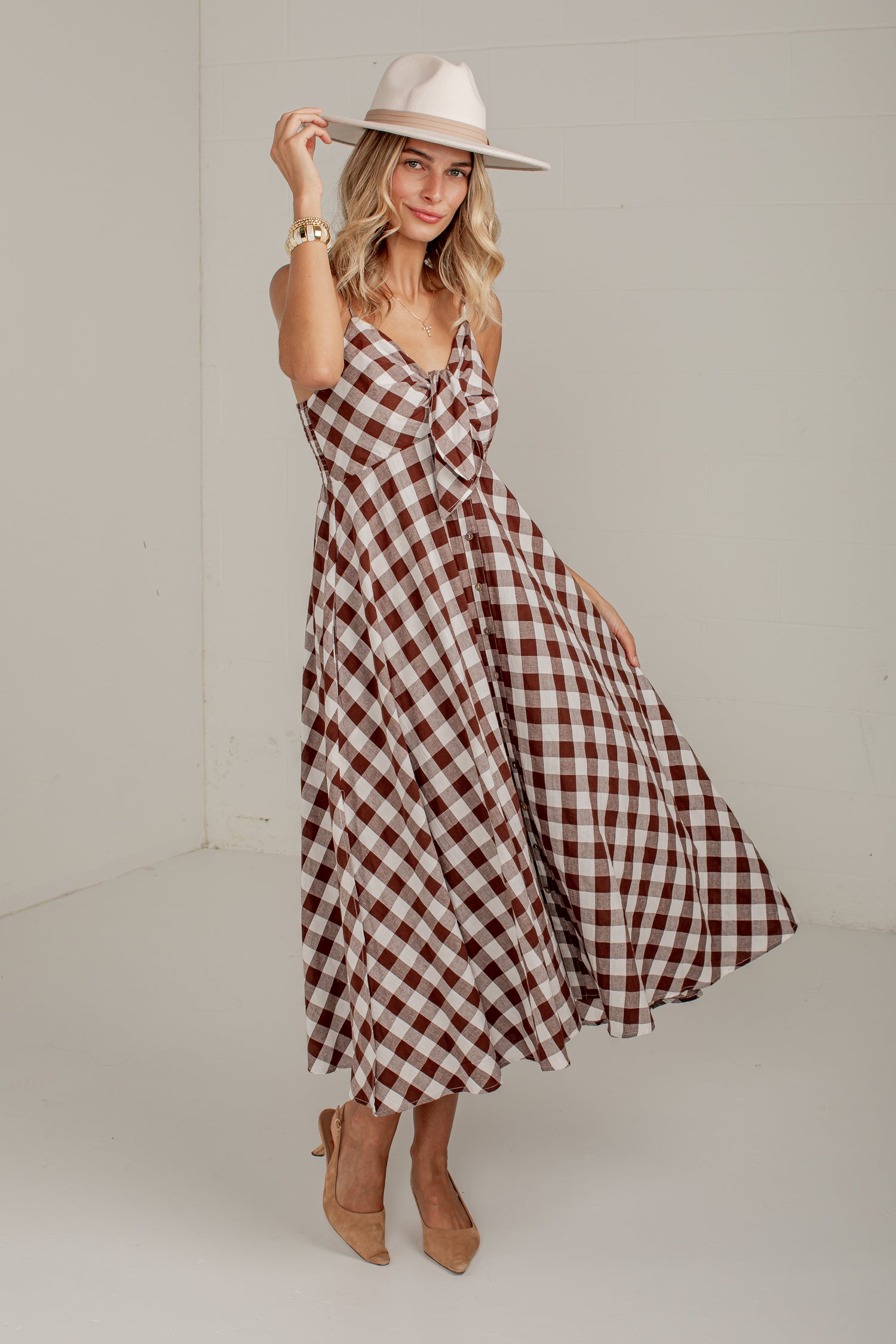 A woman modeling a brown and white gingham midi dress with a knotted bodice, styled with a wide-brimmed cream hat and tan heels in a neutral studio setting.