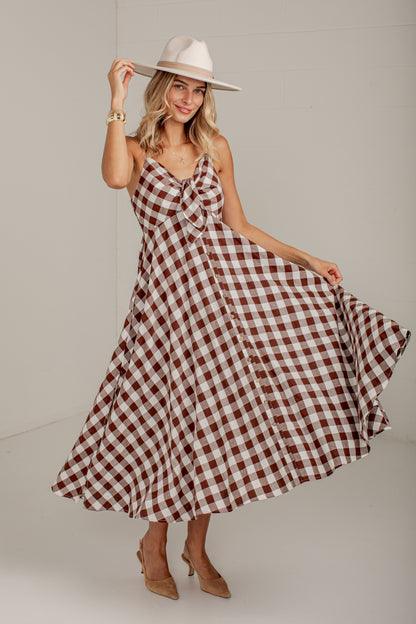 A woman modeling a brown and white gingham midi dress with spaghetti straps and a sweetheart neckline, paired with a cream wide-brim hat and tan heels, set against a plain light gray studio background.