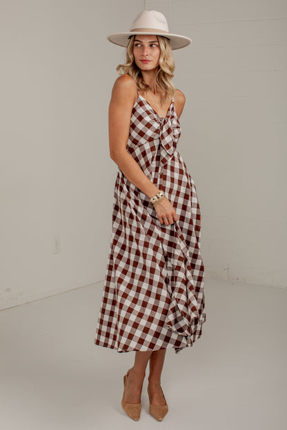 A woman modeling a brown and white plaid midi dress with a wrap-style V-neck and ruffled skirt details, paired with a cream wide-brim hat and tan heels, in a minimalist studio setting.