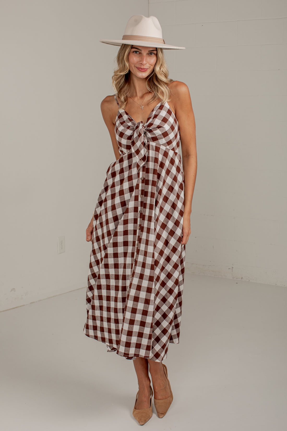 A woman modeling a sleeveless gingham midi dress with a brown and cream check pattern, a knotted neckline, and a tie waist, styled with a wide-brimmed cream hat and tan heels.