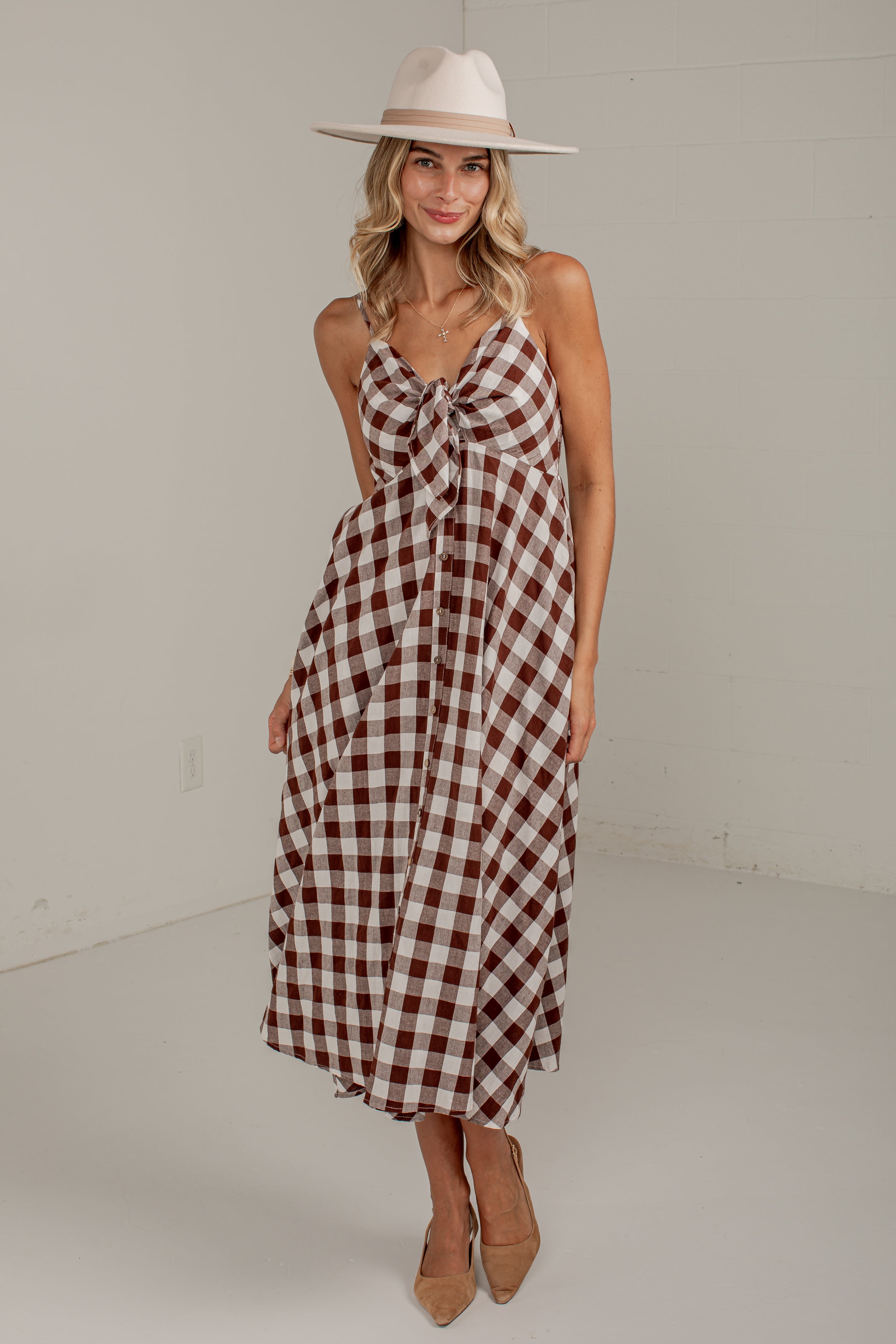 A woman modeling a sleeveless gingham midi dress with a brown and cream check pattern, a knotted neckline, and a tie waist, styled with a wide-brimmed cream hat and tan heels.