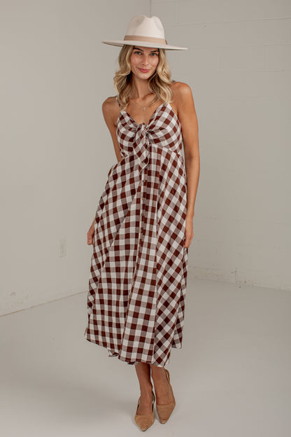 A woman modeling a sleeveless gingham midi dress with a brown and cream check pattern, a knotted neckline, and a tie waist, styled with a wide-brimmed cream hat and tan heels.