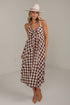 A woman modeling a sleeveless gingham midi dress with a brown and cream check pattern, a knotted neckline, and a tie waist, styled with a wide-brimmed cream hat and tan heels.