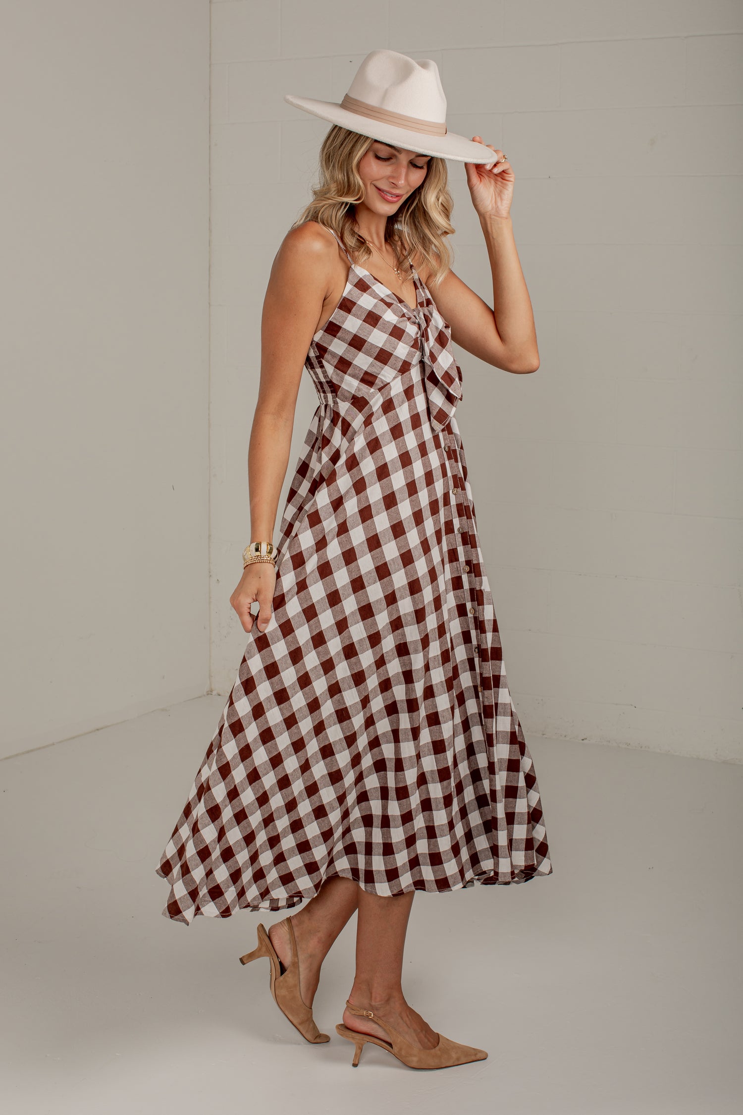 A woman modeling a brown and white gingham midi dress with spaghetti straps and a tie detail at the bust, styled with a wide-brimmed cream hat and tan slingback heels against a plain light gray studio background.