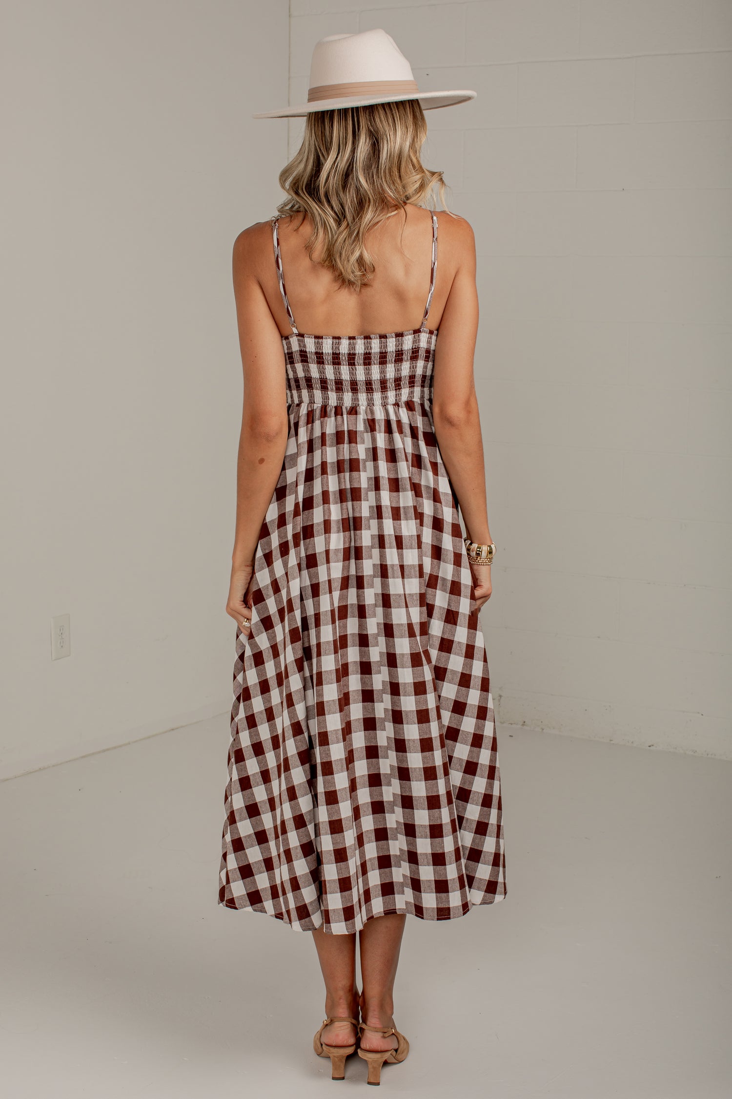 A woman viewed from behind wearing a brown and white gingham midi dress with thin spaghetti straps and a wide-brimmed beige hat, standing in a minimalist studio setting.