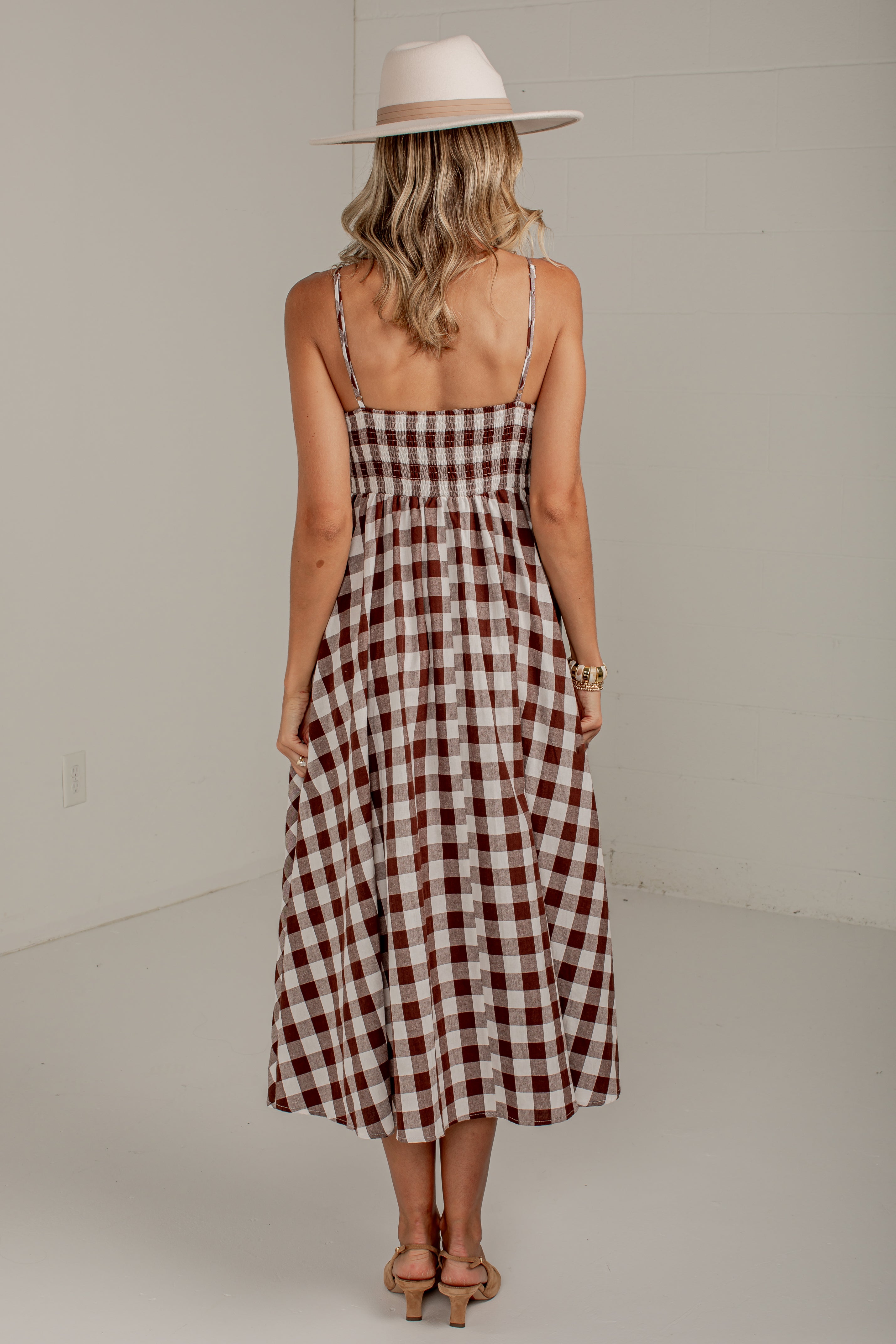 A woman viewed from behind wearing a brown and white gingham midi dress with thin spaghetti straps and a wide-brimmed beige hat, standing in a minimalist studio setting.