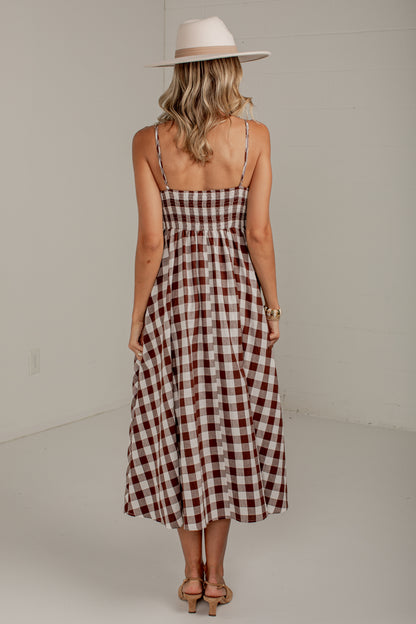 A woman viewed from behind wearing a brown and white gingham midi dress with thin spaghetti straps and a wide-brimmed beige hat, standing in a minimalist studio setting.