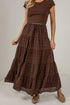 A woman wearing a brown tiered maxi skirt with a subtle plaid pattern, paired with a matching brown short-sleeve crop top and brown flat shoes, standing against a light gray wall.