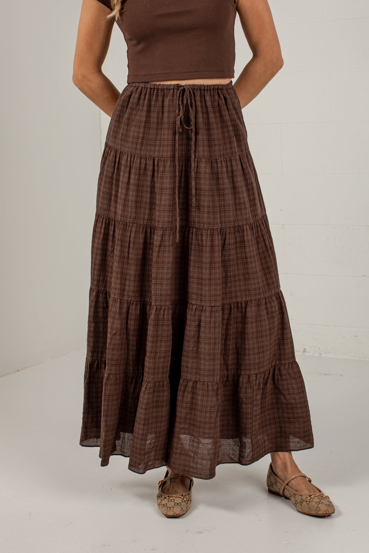 A woman wearing a brown tiered maxi skirt with a subtle plaid pattern, paired with a matching brown crop top and brown flat shoes, standing against a white wall.