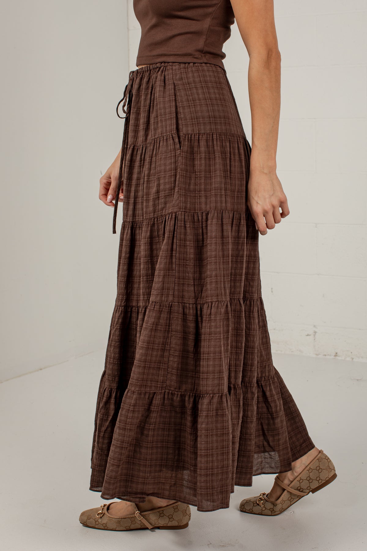 A model wearing a brown tiered maxi skirt with a subtle plaid pattern, paired with a matching brown top and brown flat shoes, standing against a white wall.