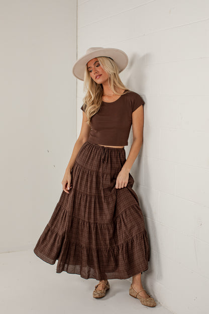 A woman wearing a brown short-sleeve crop top and a matching brown tiered maxi skirt, styled with a beige wide-brim hat and tan flat shoes, leaning against a white wall.