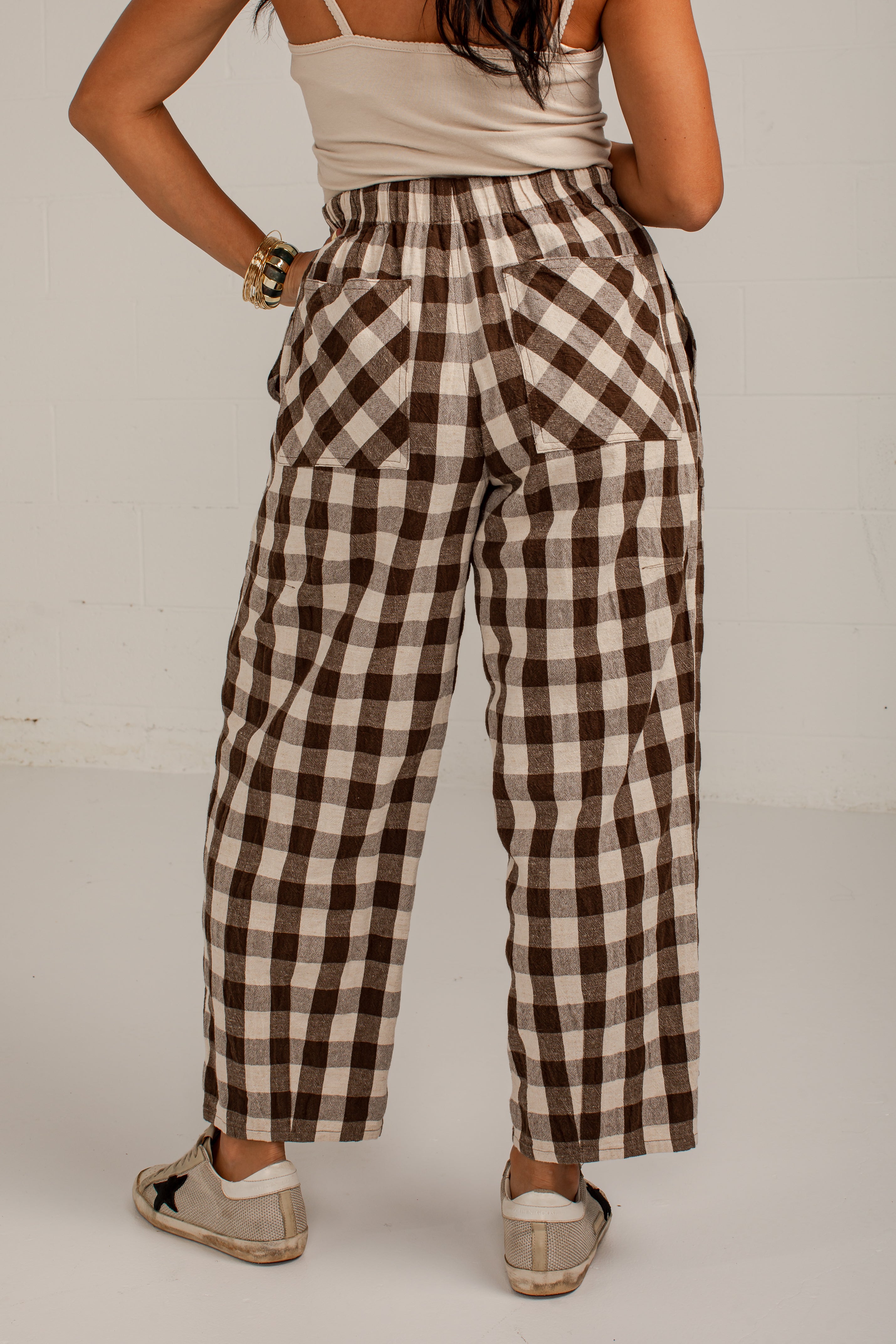 A person shown from the back wearing brown and beige gingham wide-leg pants, a cream camisole top, and beige slip-on shoes against a plain light gray studio background.