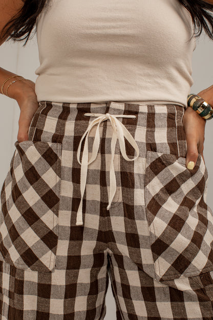 A close-up of a person wearing brown and beige buffalo plaid drawstring pants with large front patch pockets, paired with a cream ribbed tank top.