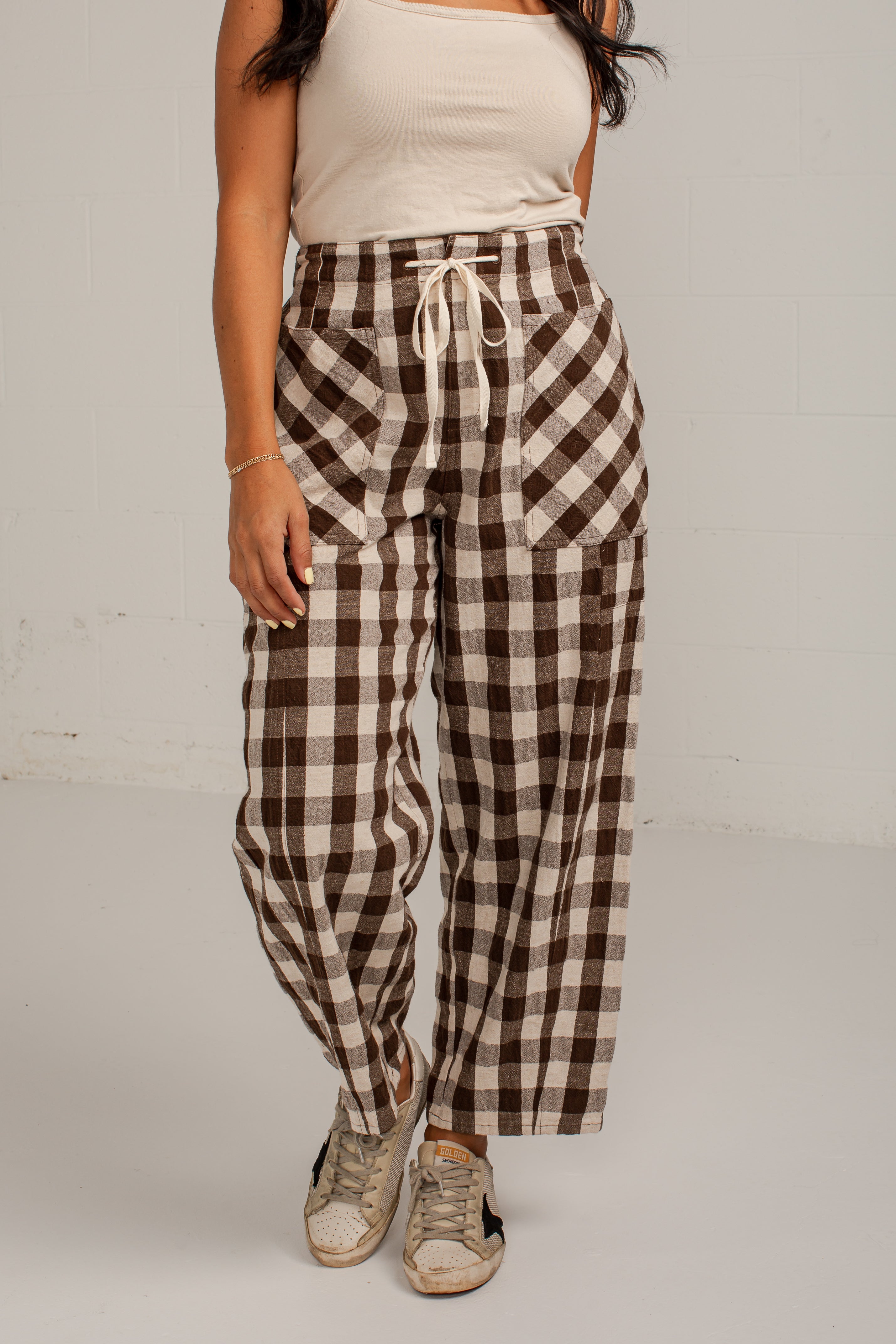 A model wearing brown and beige gingham wide-leg pants with a drawstring waist, paired with a cream tank top and beige sneakers, standing against a white wall.