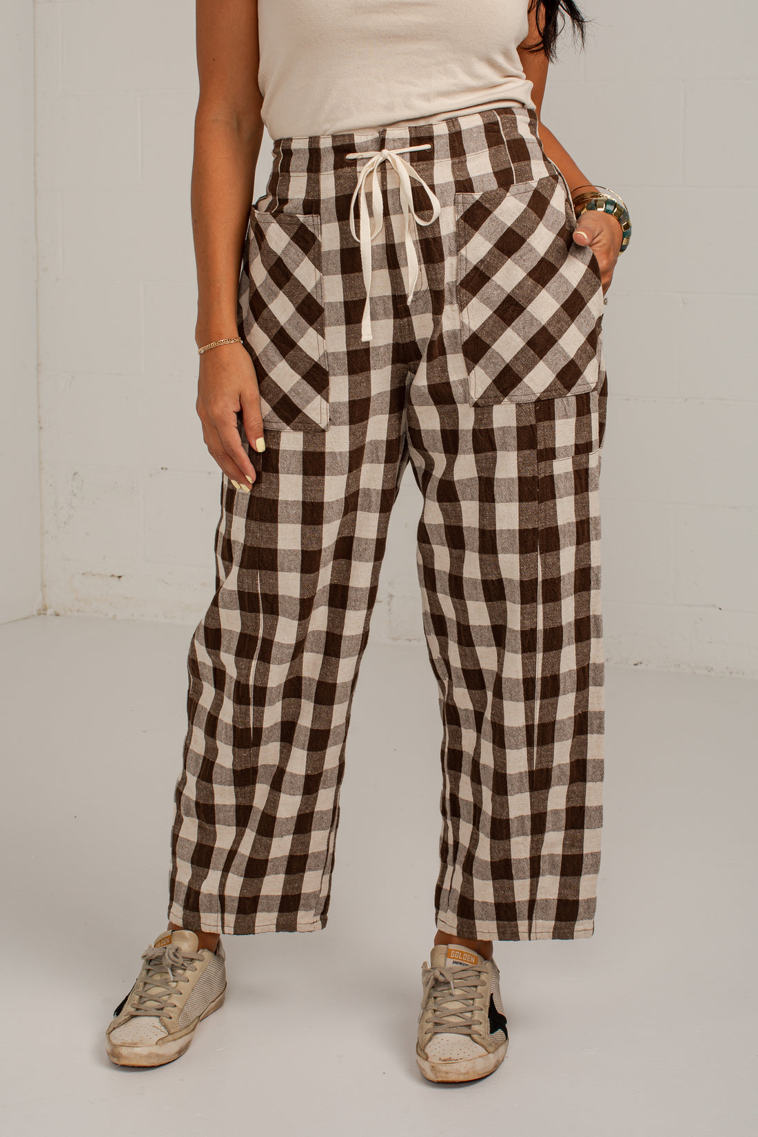 A person wearing brown and beige buffalo plaid cropped pants with a white drawstring waist, paired with a cream top and beige sneakers, standing against a white wall.