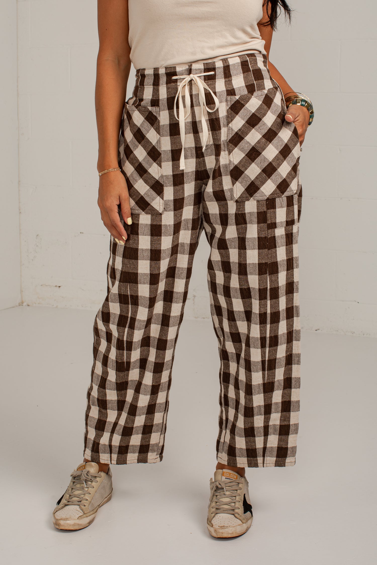 A person wearing brown and beige buffalo plaid cropped pants with a white drawstring waist, paired with a cream top and beige sneakers, standing against a white wall.