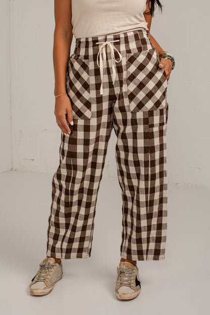 A person wearing brown and beige buffalo plaid cropped pants with a white drawstring waist, paired with a cream top and beige sneakers, standing against a white wall.