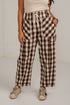 A person wearing brown and beige buffalo plaid cropped pants with a white drawstring waist, paired with a cream top and beige sneakers, standing against a white wall.