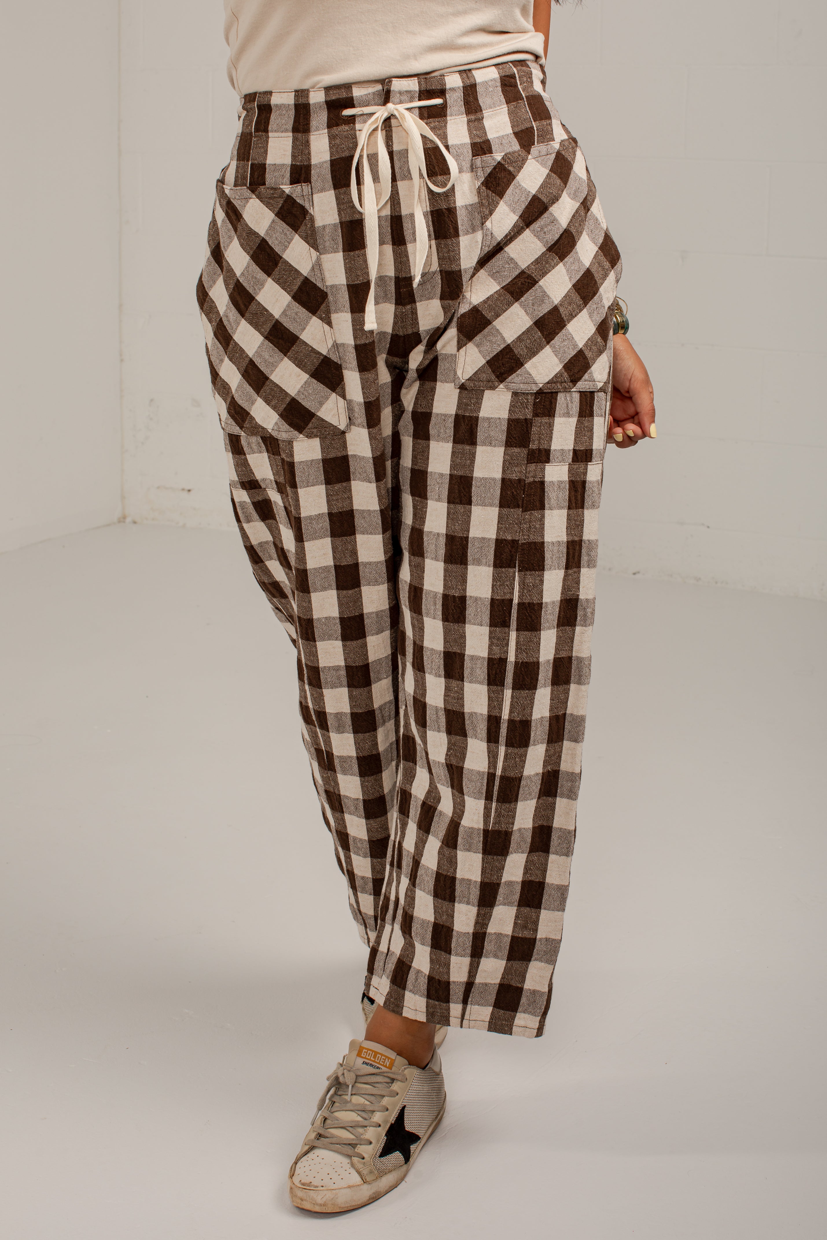 A person wearing brown and beige gingham check cropped pants with a cream drawstring waist, paired with a cream top and distressed sneakers, photographed in a studio setting.