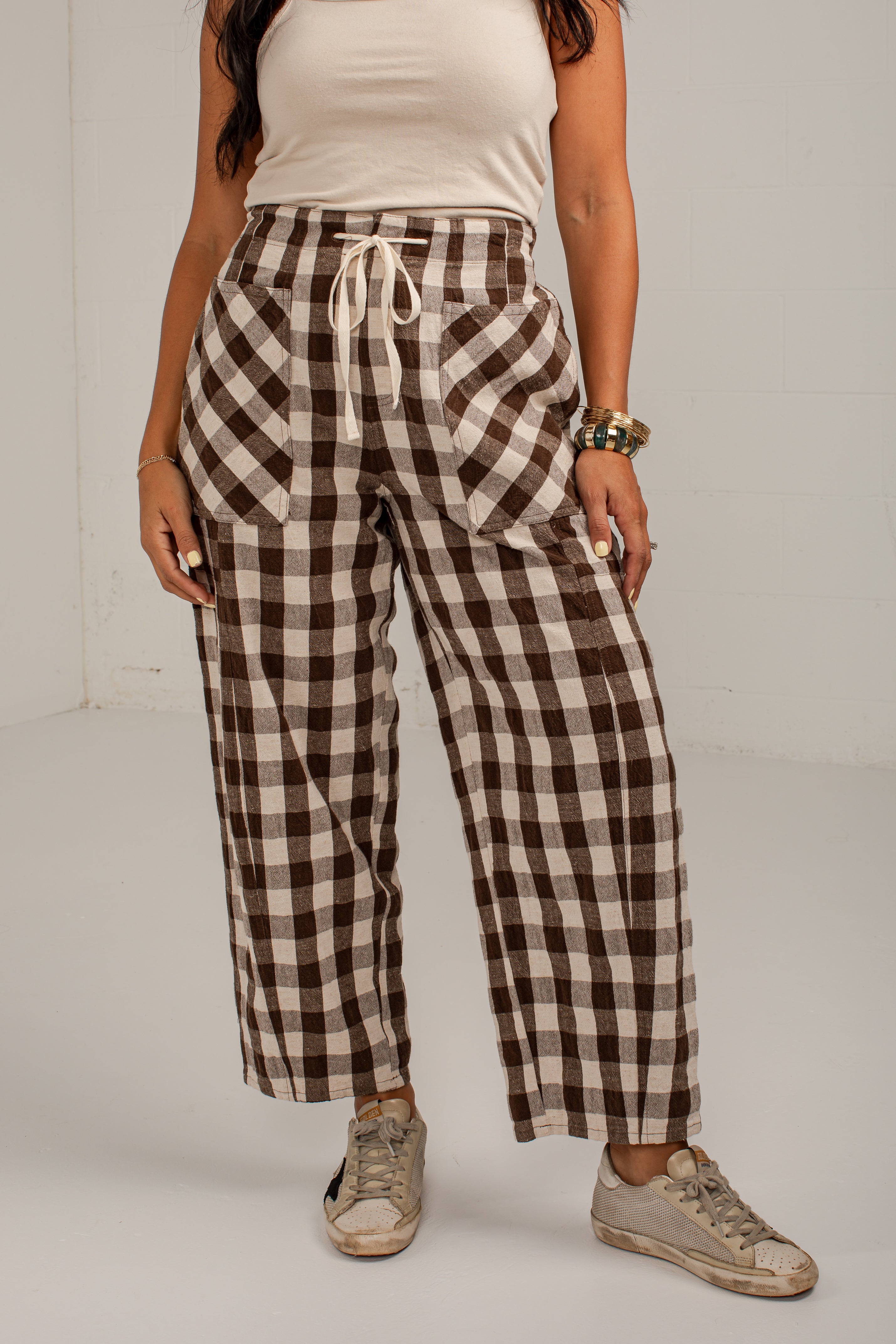 A person wearing brown and beige checkered wide-leg cropped pants with a drawstring waist, paired with a cream tank top and beige sneakers, standing against a plain light gray studio background.