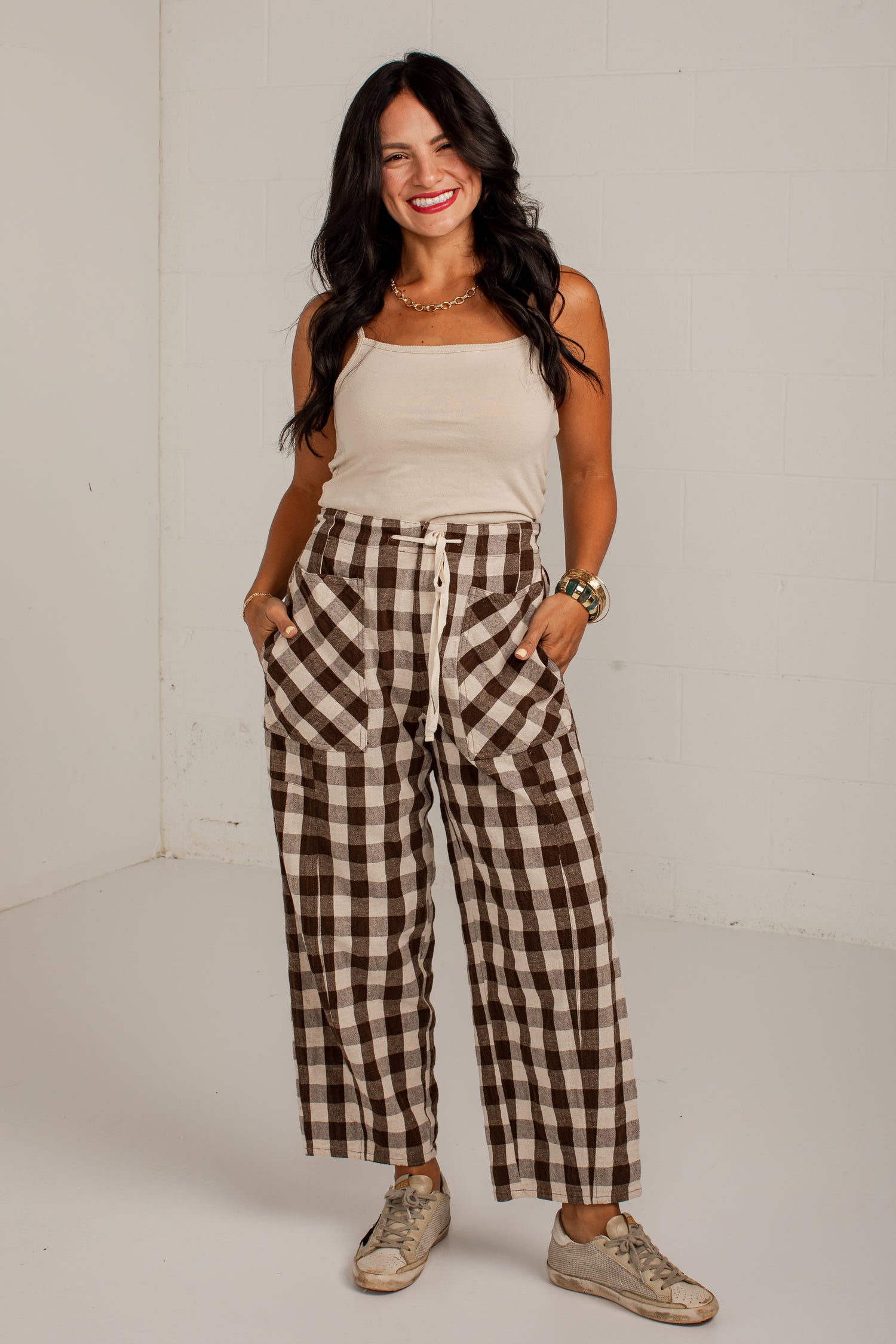 A woman modeling a pair of brown and beige plaid wide-leg pants with a drawstring waist, paired with a cream tank top and beige sneakers, standing against a white studio wall.