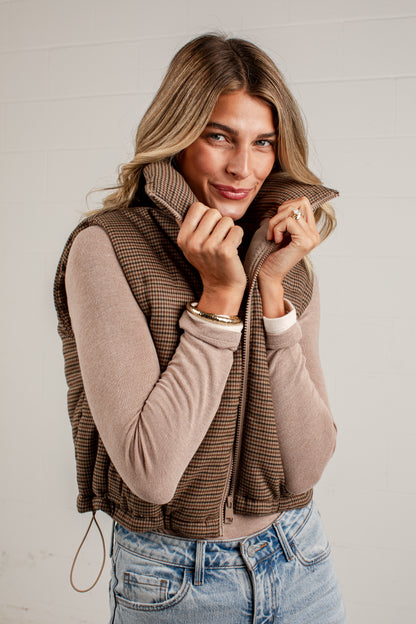 A woman wearing a brown and beige houndstooth zip-up vest over a beige long-sleeve top and blue jeans, posing against a white paneled wall.