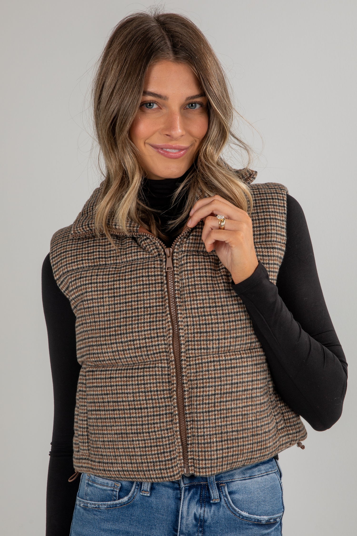 Woman wearing a plaid vest over a black long-sleeve shirt and blue jeans on a gray background