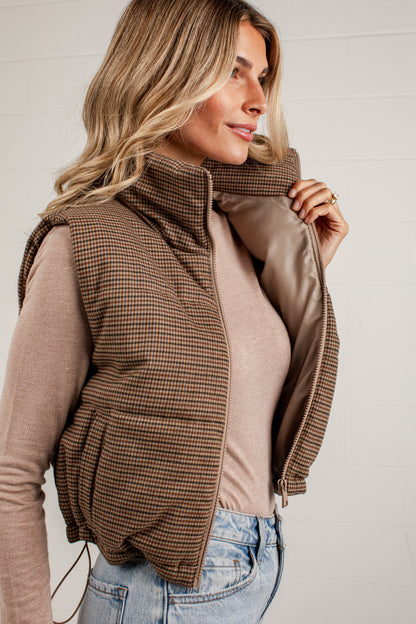 A woman wearing a brown and beige houndstooth puffer vest over a beige long-sleeve top and blue jeans, shown in a close-up profile view against a light gray paneled wall.