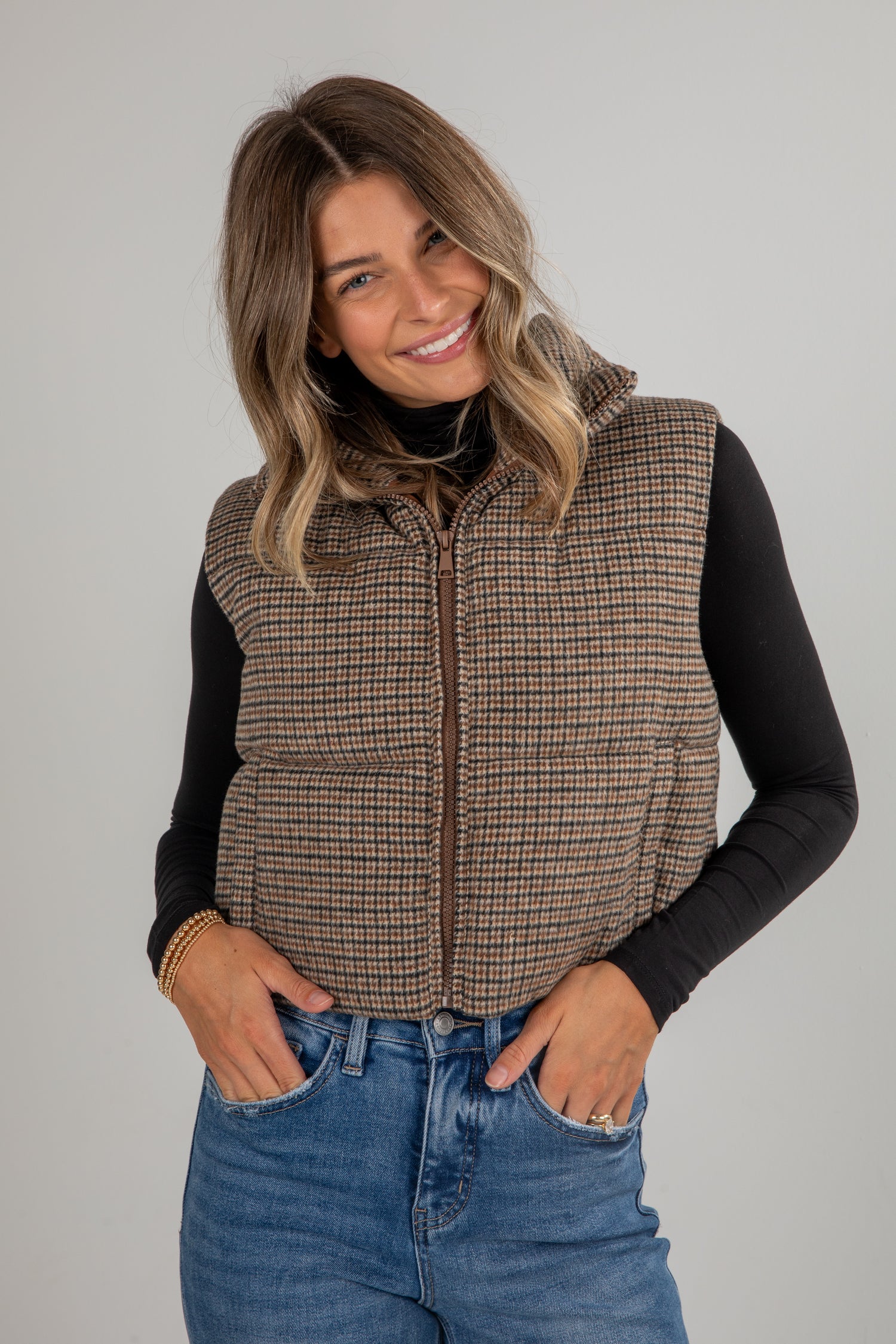 Woman wearing a plaid vest over a black long-sleeve shirt and blue jeans on a gray background