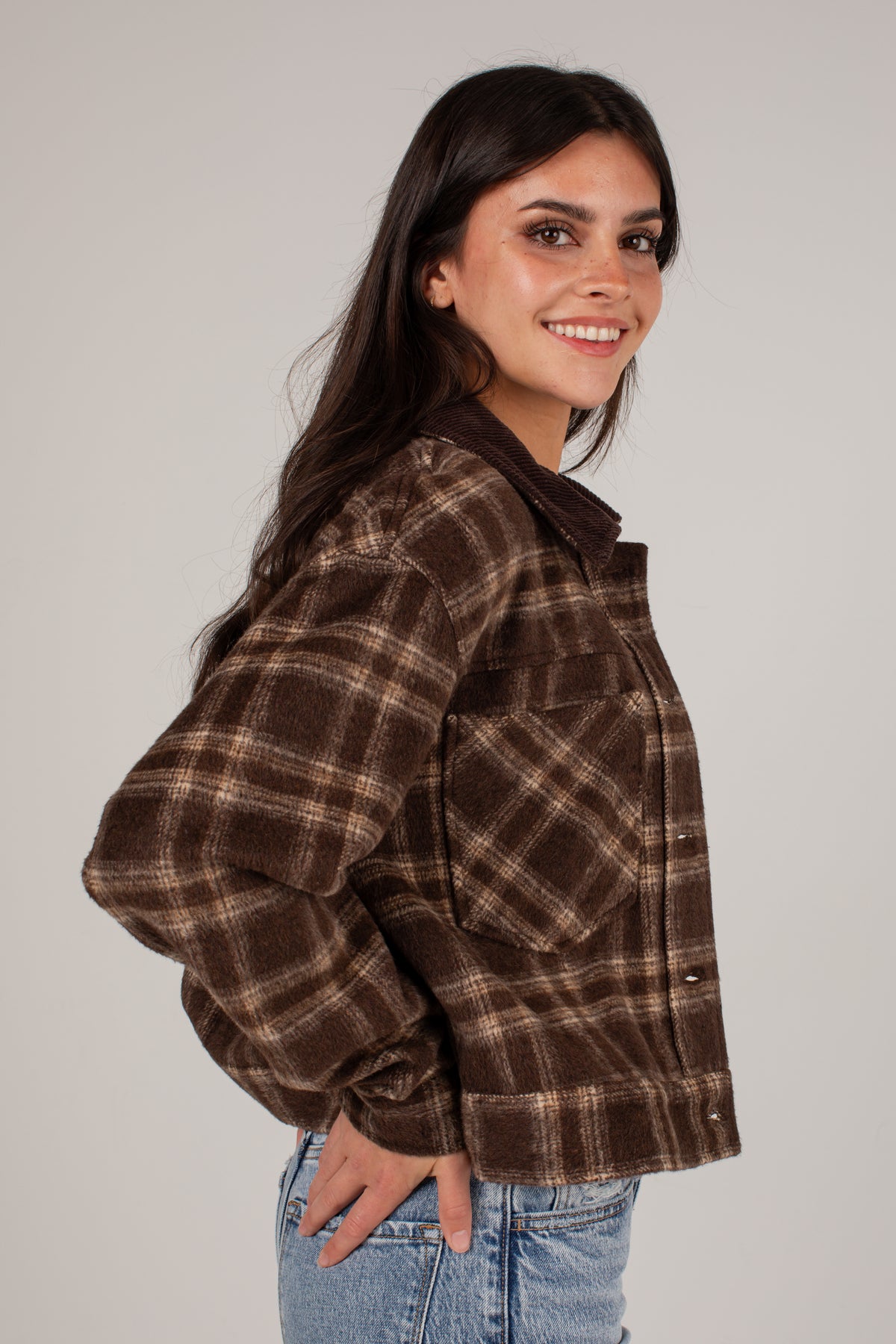 A woman wearing a brown and beige plaid flannel jacket, paired with light blue jeans, photographed against a solid light gray background.