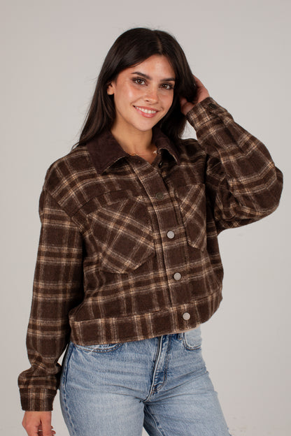 A woman wearing a brown and beige plaid cropped flannel shirt jacket with a solid brown corduroy collar, paired with light blue jeans, against a plain light gray background.