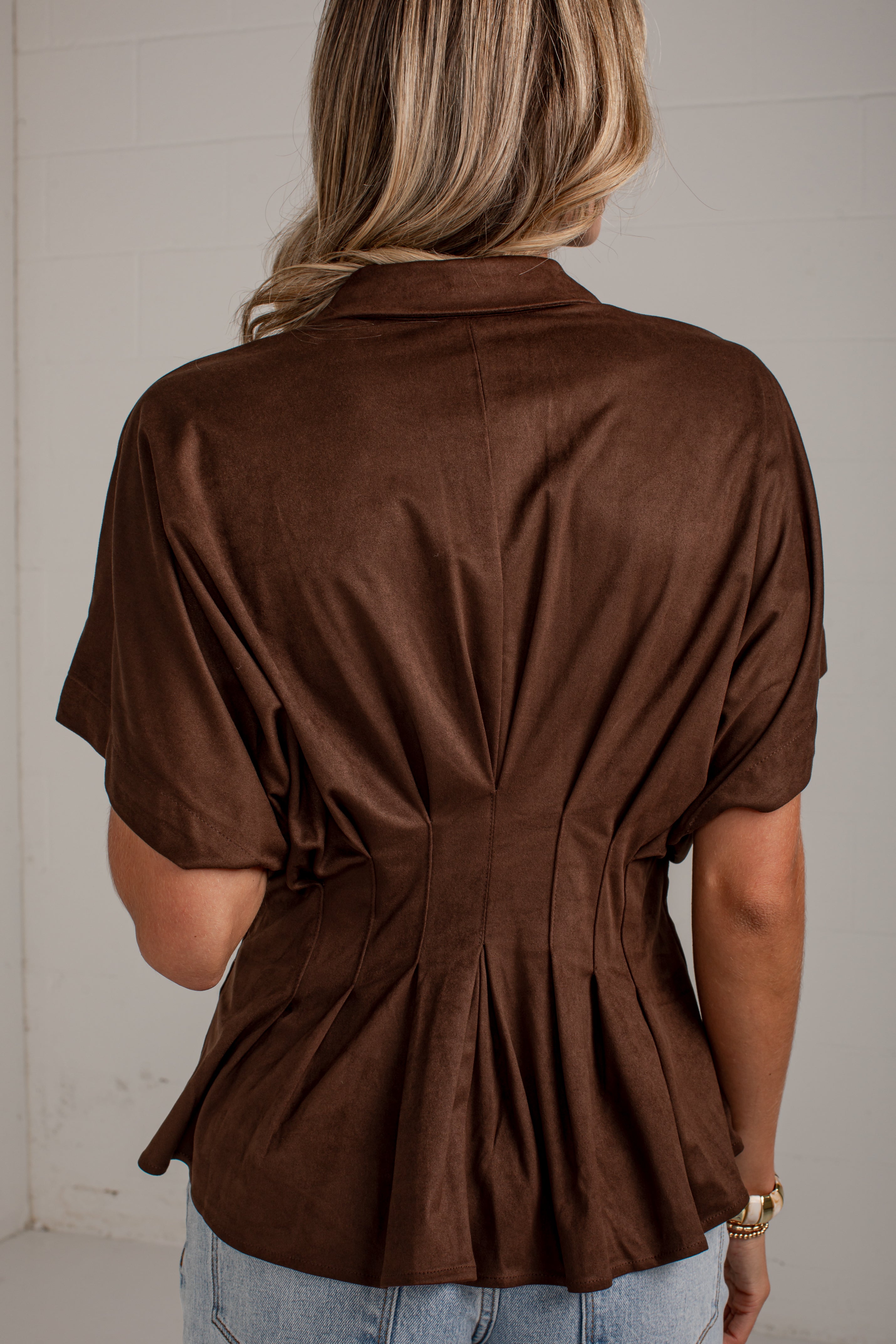 A woman viewed from behind wearing a brown short-sleeve collared blouse with gathered detailing at the back and light blue jeans, standing against a light neutral wall.