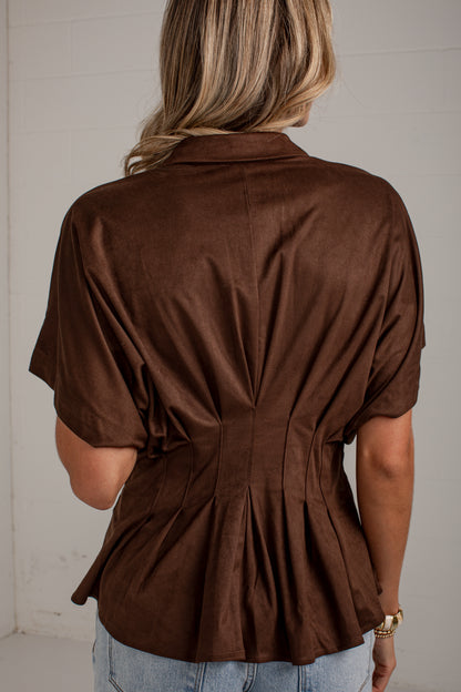 A woman viewed from behind wearing a brown short-sleeve collared blouse with gathered detailing at the back and light blue jeans, standing against a light neutral wall.