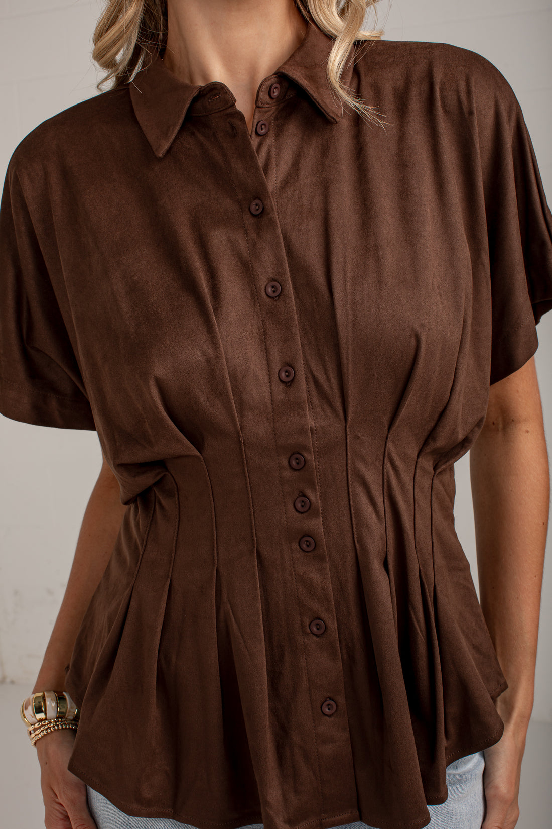 A woman wearing a brown short-sleeve button-up shirt with a peplum waist and gathered side details, shown from the shoulders to the hips against a plain light background.