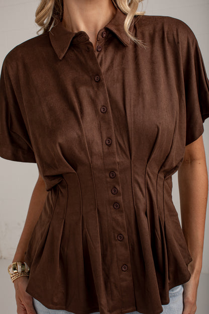 A woman wearing a brown short-sleeve button-up shirt with a peplum waist and gathered side details, shown from the shoulders to the hips against a plain light background.