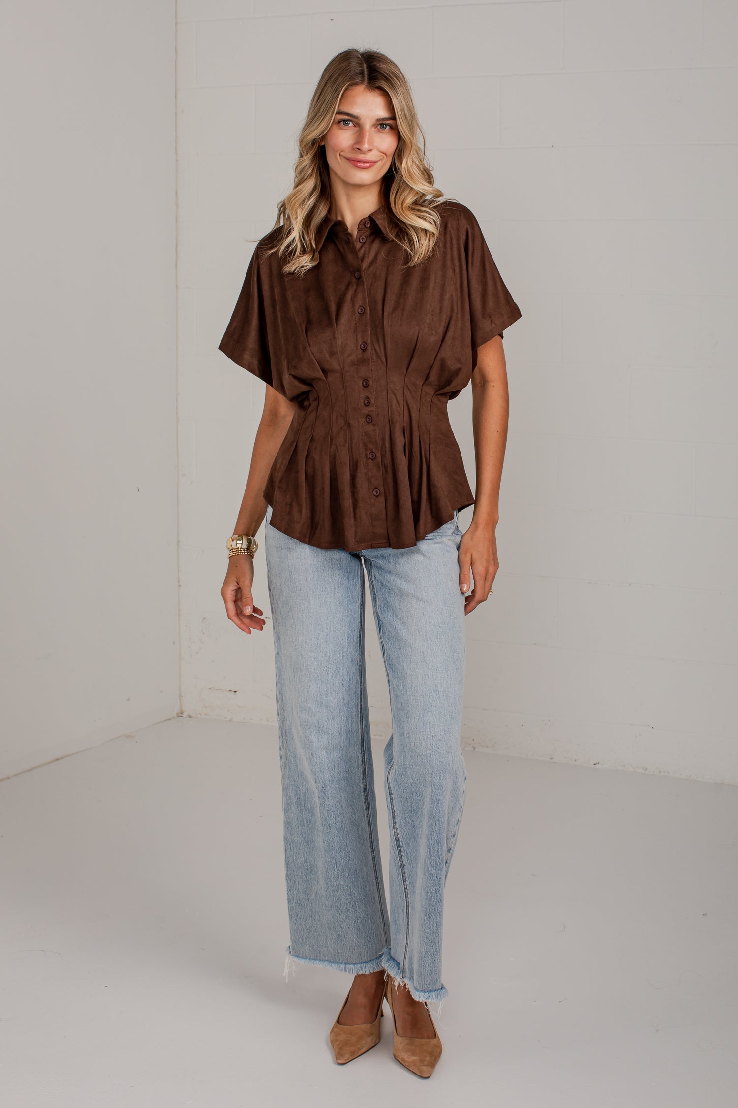 A woman modeling a brown short-sleeve button-up peplum blouse with light-wash wide-leg jeans and tan pointed-toe heels, standing against a plain white studio wall.